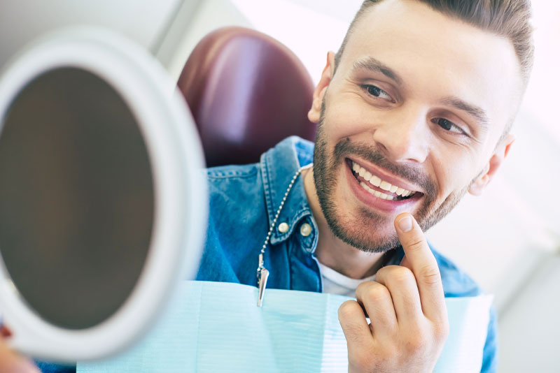 a picture of a dental patient smiling into a hand held mirror and pointing at his new smile because he was just treated with customized dental veneers.
