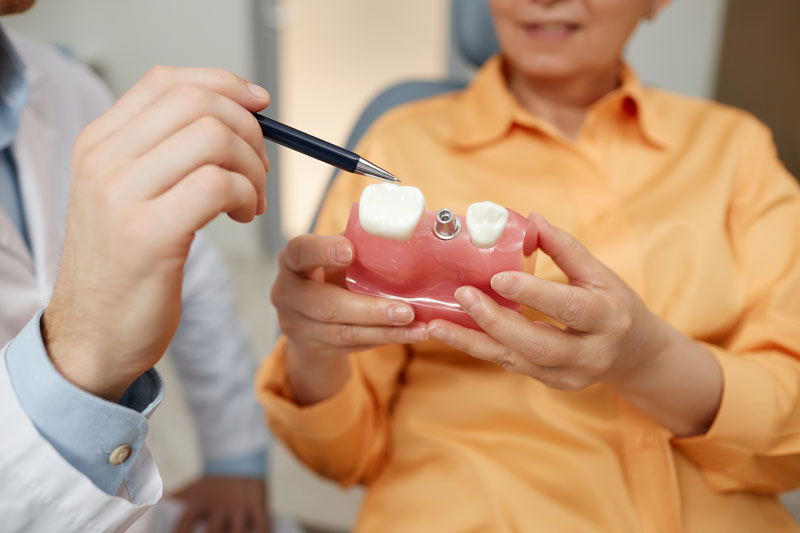 Closeup of unrecognizable dentist pointing at tooth model while explaining dental implant surgery to patient in clinic